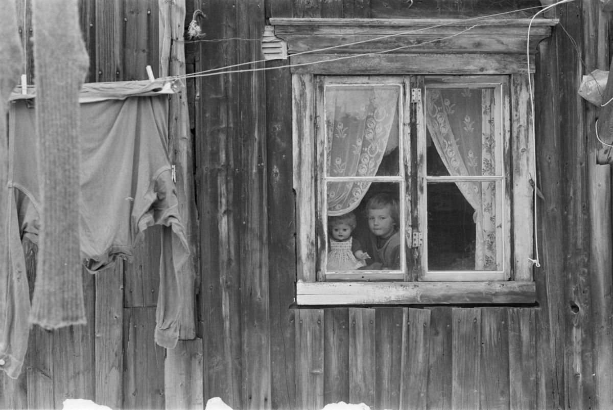 Et barn med en dukke titter ut av et vindu.