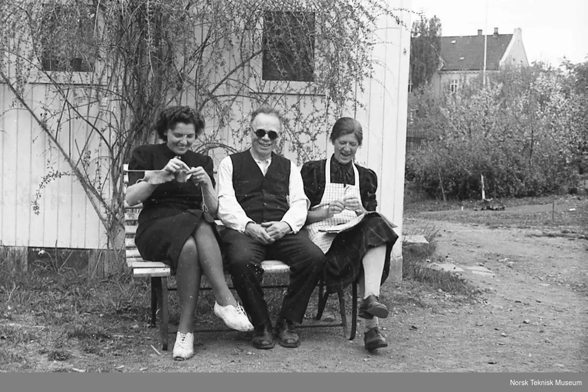 Annie Halvorsen strikker, Hans Halvorsen sitter og Beth Marie Halvorsen leser avisen på benken i hagen, Hasleveien 42, våren 1940