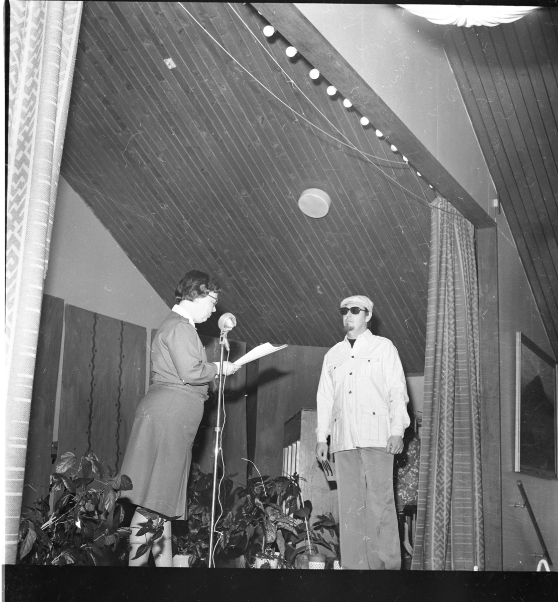 En kvinna och en man står på en scen, möjligen i samband med modevisning. Kvinnan står vid en mikrofon och läser i ett papper. Hon bär Lottakårens uniform.