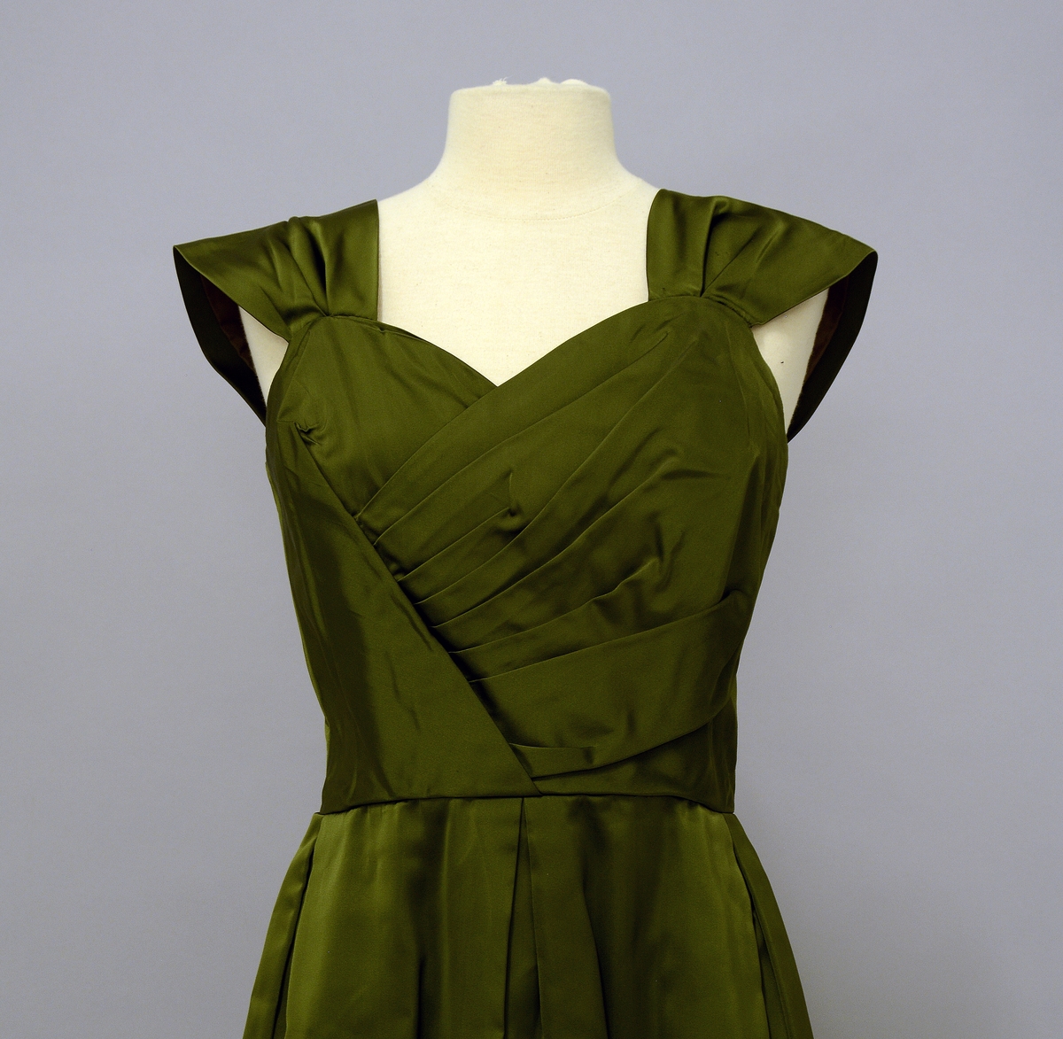Selskapskjole med brede stropper på skulderene. Foran har kjolen flere legg på skrå som dekor. Kjolen kan lukkes med glidelås bak. Skjøret er lagt i brede folder foran og bak. Tøyet er av grønn silkesateng. Det tilhører en grønn stola til kjolen: TGM.2020:046.B
