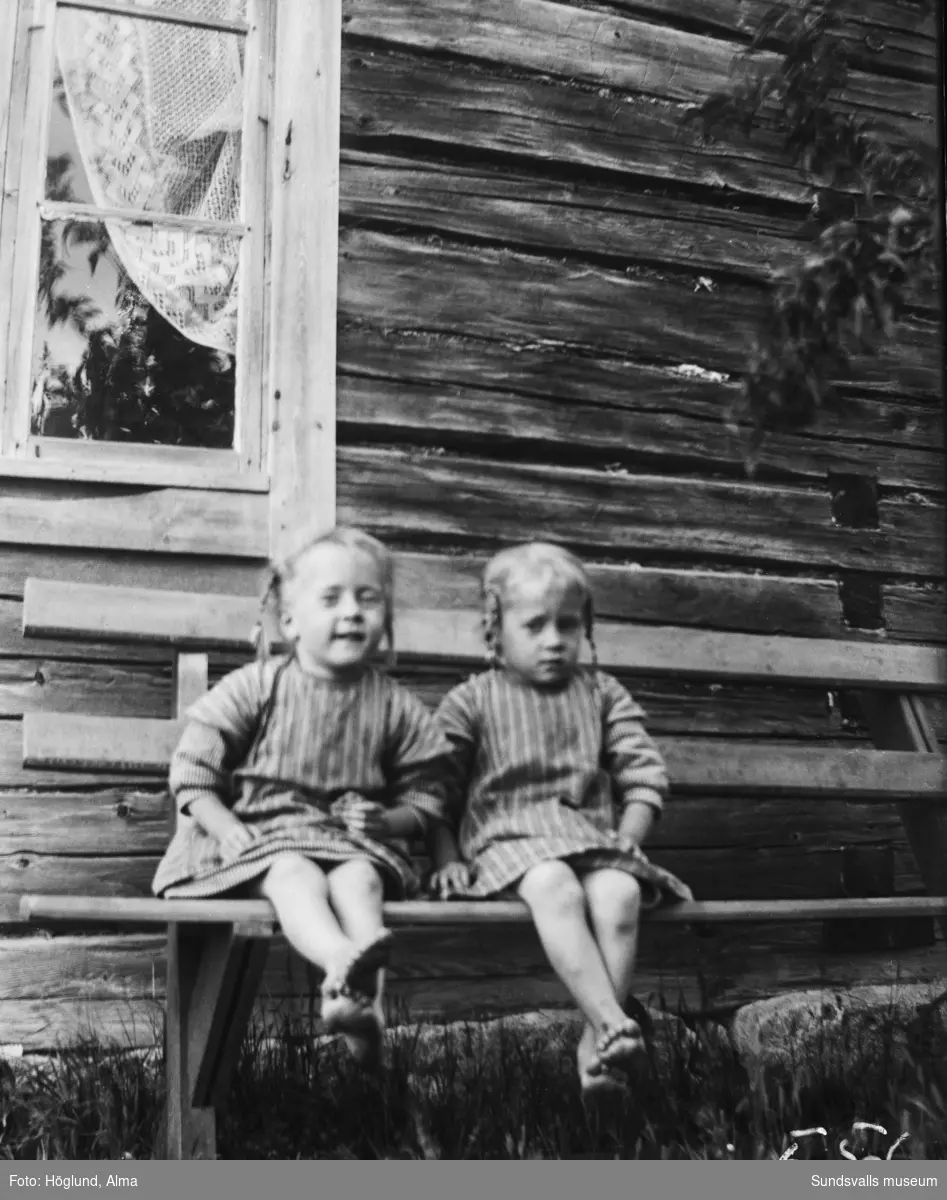Tvillingsystrarna Ingrid och Irma Höglund på en bänk invid ett timrat hus.