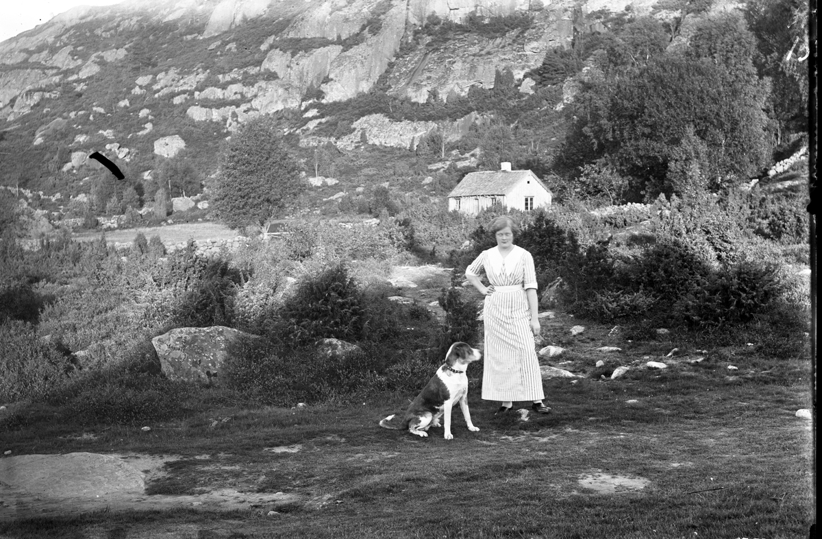 En kvinna i randig klänning står med en stor hund i naturen. I bakgrunden reser sig en hög bergvägg och vid dess fot ligger ett hus ligger rakt bakom kvinnan.