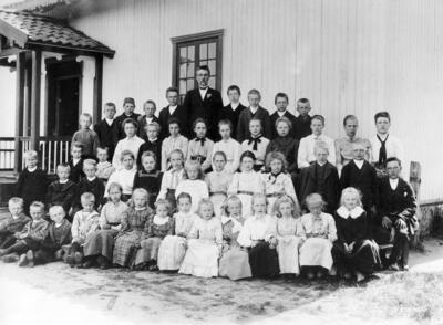 Fotografi i svart-hvitt av en skoleklasse og lærer Simensen. Elevene og læreren blir fotografert utenfor Skinnarbøl skole, som nå er skolemuseum.