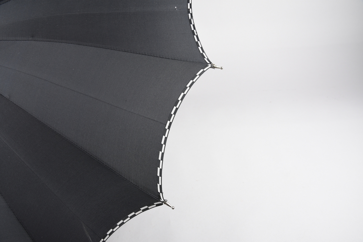Paraply med 16 metallspilar. Stong i tre. Trekt med stoff i kunstfiber, ensfarga svart med dekor i sølv/svart rutemønster. Handtak i plast med hempe i tekstil til å ta inn på handa. Hempe for lukking av paraplyen.