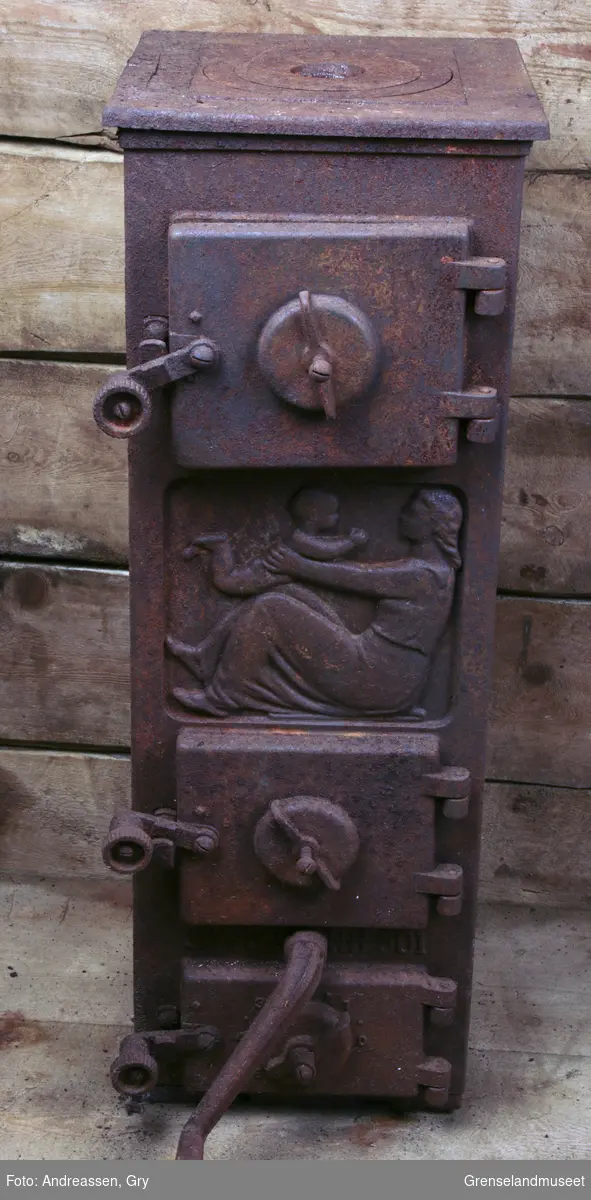 Ovn, JØTUL 501, rusten og falleferdig. Et panel har bilde av en kvinne sittende på bakken, som løfter et barn opp med armer og knær. Den har tre dører.