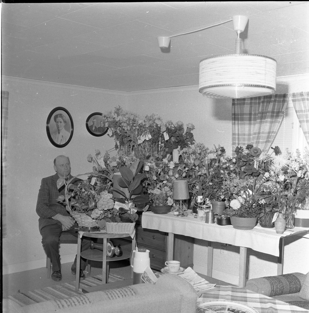 En man sitter vid ett bord fullt av blomsterbuketter, det är mannens 50-årsdag. På väggen bakom honom hänger två äldre ovala fotografier av en kvinna respektive kvinna och man