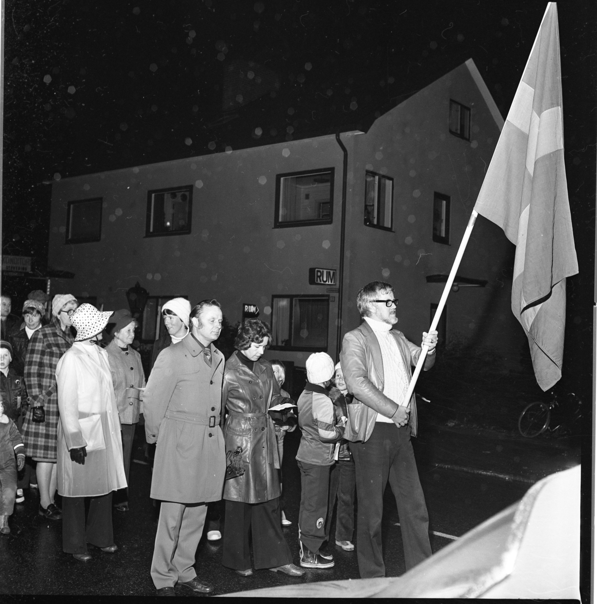 Vuxna och enstaka barn går i ett led efter en fanbärare med svensk flagga. Grännabor demonstrerar för att få en sporthall till Gränna. Det är oktober, mörkt och blött. Mannen i rock är Allan Svensson.