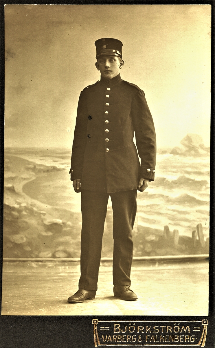 Okänd ung man i någon sorts uniform, fotograferad i helfigur framför en landskapsfond, fotograferad i Varberg. (Se även bild nr F9142)