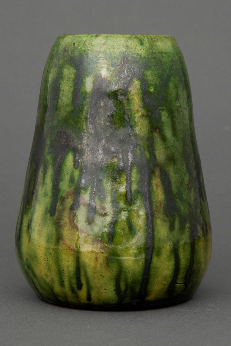 Liten vase i dreid leirgods. Vasen har pæreform og er glasert med grønn flyteglasur og rennende svart jernglasur.