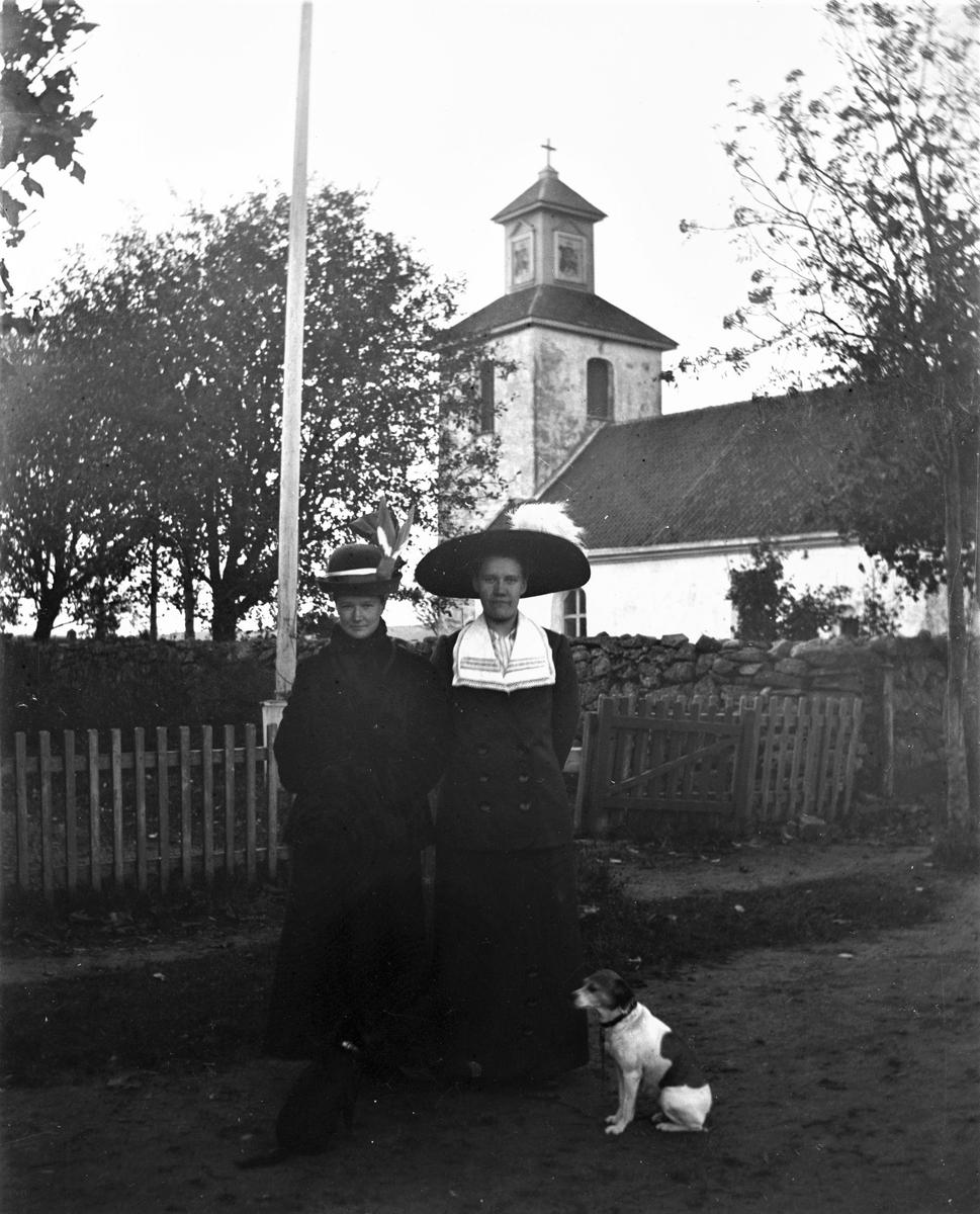 Två hattklädda unga kvinnor med var sin liten hund står utanför kyrkan i Karl Gustav. Bakom dem står grinden öppen i trästaketet som hägnar en trädgård med flaggstång. (Kvinnan till höger finns även på bild nr GEB166)