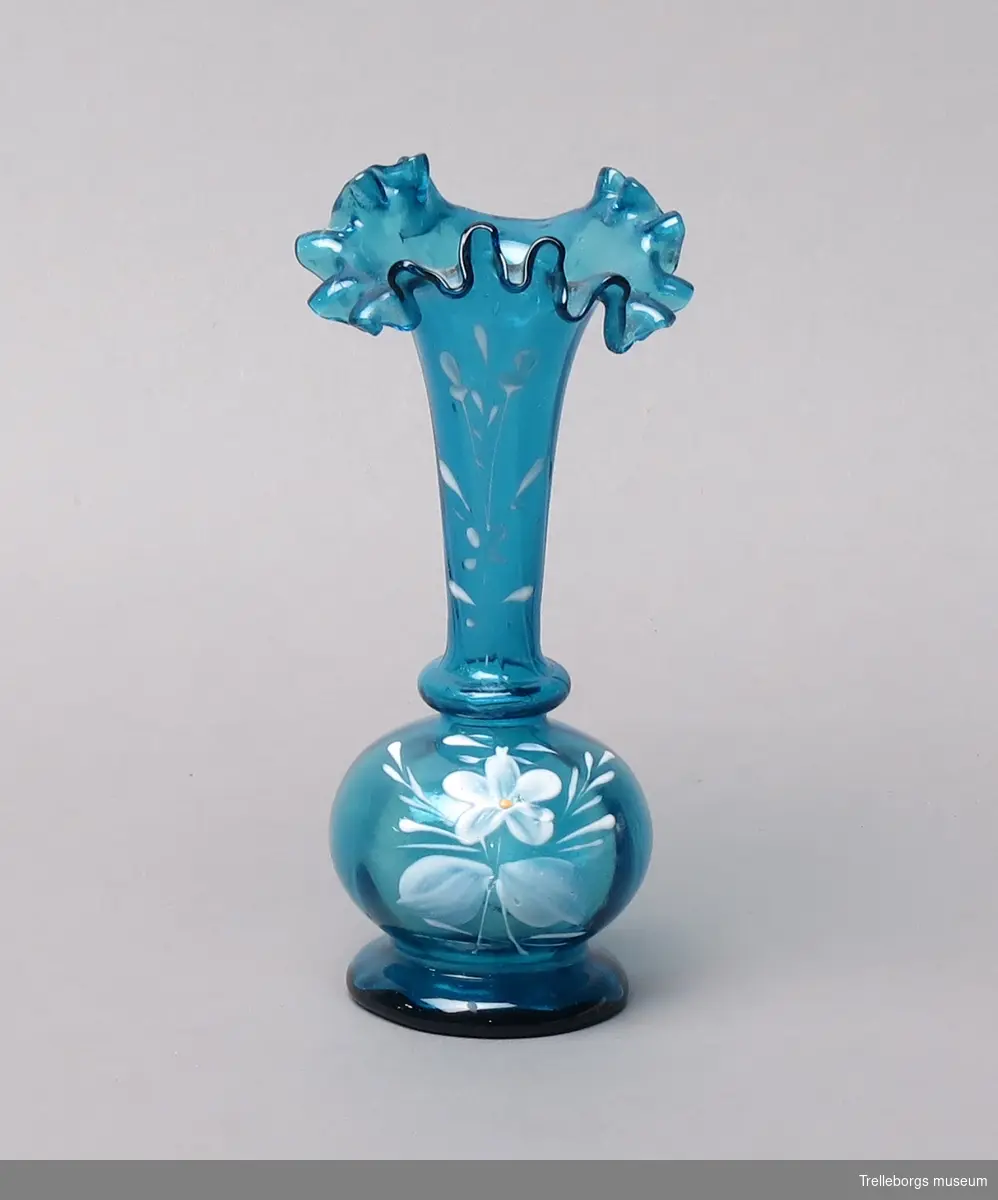 Vas med påmålad dekor, med en liten fot (diameter 4,5 cm) och ovanpå den en rund del. Från den går vasen upp i en strutform, som vrider sig svagt, med veckade kanter.