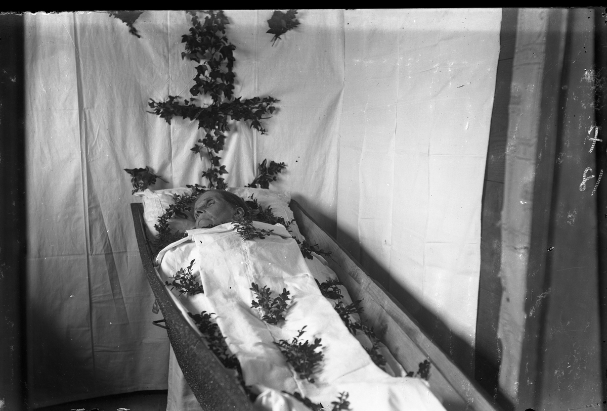 En gammal kvinna "ligger lik" i hemmet. Kistan är placerad i ett hörn där lakan hängts upp på väggarna. I kistan ligger små kvistar av buxbom utspridda och fäst på lakanet ovanför huvudgärden sitter ett kors av murgröna,

Ända sedan hednisk tid, och fram tills det i Sverige förbjöds 1686, höll man likvaka/likvakt natten till begravningen av en död (gravöl hölls däremot efter begravningen för att hedra och minnas den döde ). En anledning till att seden då förbjöds i kyrkolagen var att vakan ofta antog karaktären av fest med sprit och dans, vilket inte gick ihop med den Lutherska moralen. Seden övergick till att man gick för att "se liket", vilket förmodligen är fallet vid denna fotografering, utan en regelrätt likvaka. Detta lever ännu kvar inom katolicismen där prominenta personer kan läggas på "lit de parade", i betydelsen paradsäng; en säng som sörjande kan paradera förbi och ta farväl av den avlidna före begravningen.