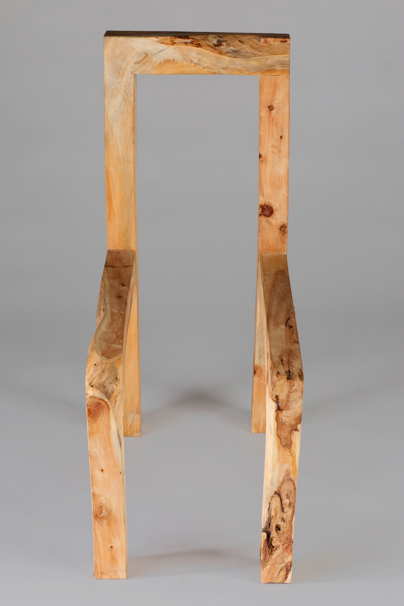 Skulpturelt objekt i tre, utformet som en stol uten sete. Høyreist rygg som består av to stolper som glir over i bakbeina, og forbundet hverandre med et toppstykke. Sarg og forbein er laget av samme treemne.