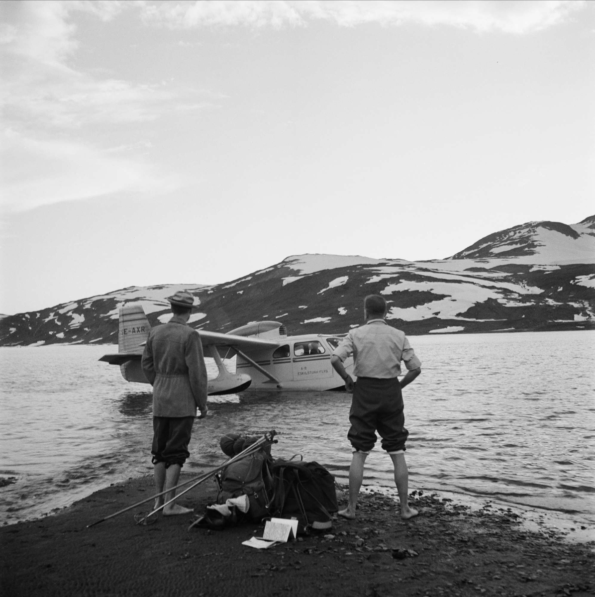 Dag Hammarskjöld och Sverker Åström på fjällvandring på Sulitelma. Två män står på stranden intill packning och utrustning. De ser ut över vattnet där ett sjöflygplan av modell Republic Seabee landat.