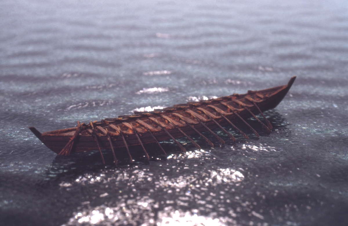 Fartygsmodell, vattenlinjemodell, hel, i "block av trä. "Nydambåten" (350 e.Kr), brunmålat skrov med höga för- och akterstävtimmer, fast sidoroder, 15 tofter, 15 par årtullar, 15 par åror. Längd i vattenlinjens 10,0 cm.Föremålets form: Vattenlinjemodell