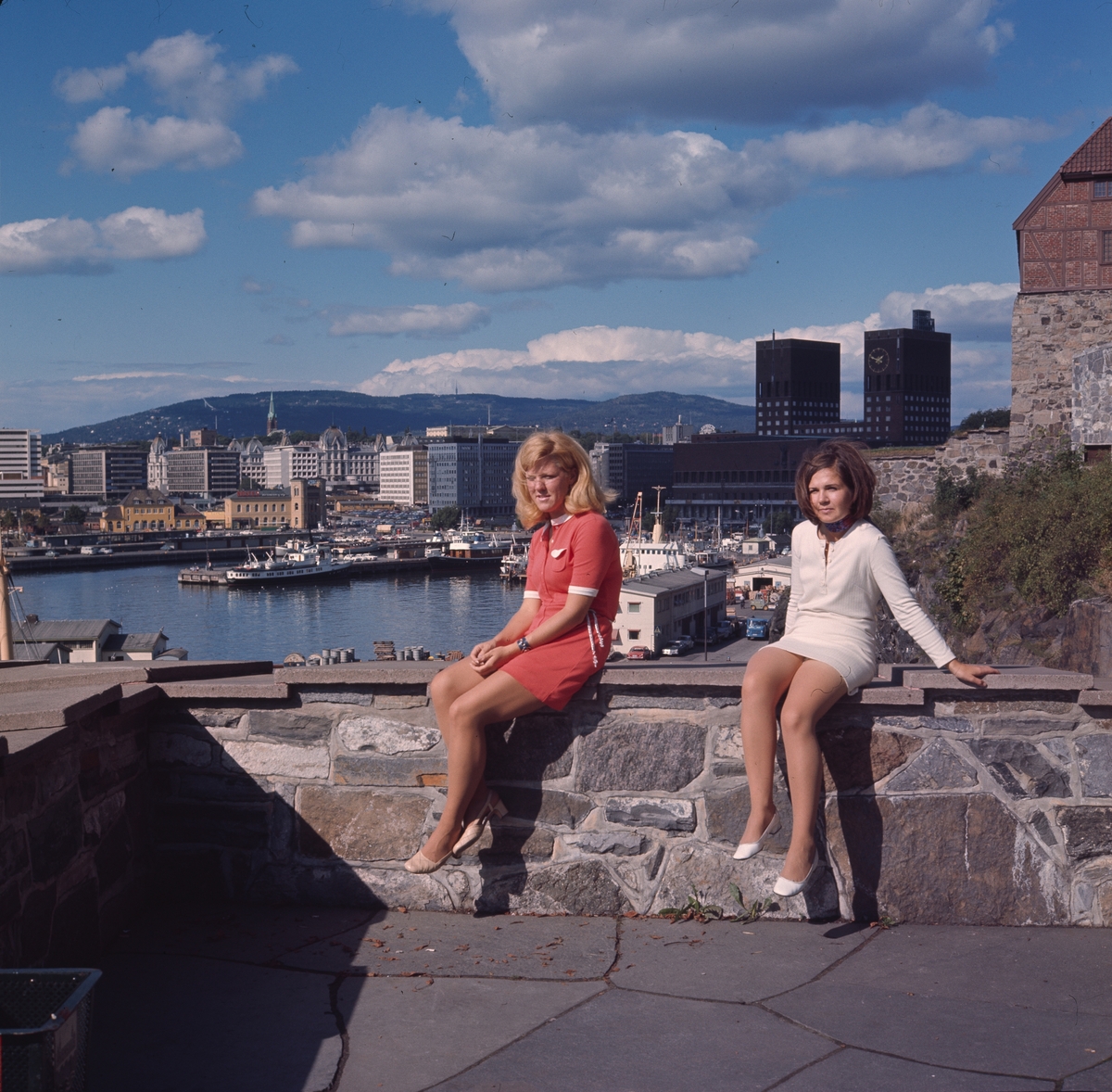 To kvinner sitter på en mur på Akershus festning i august 1969. De er kledd i korte, tidstypiske sommerkjoler. Utsikt til Rådhuset, Vika og Vestbanen.