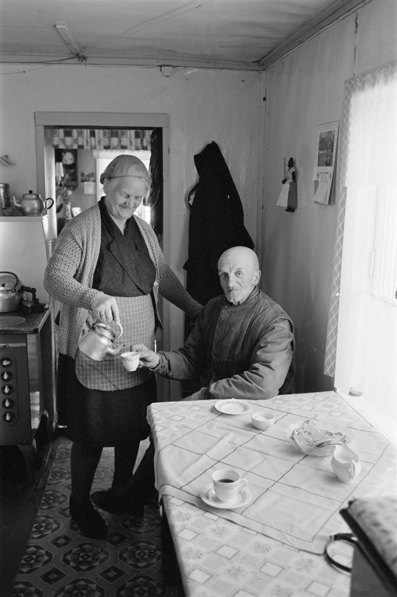 En kvinne skjenker en kopp kaffe til en mann sittende ved et bord i en stue på Røros.