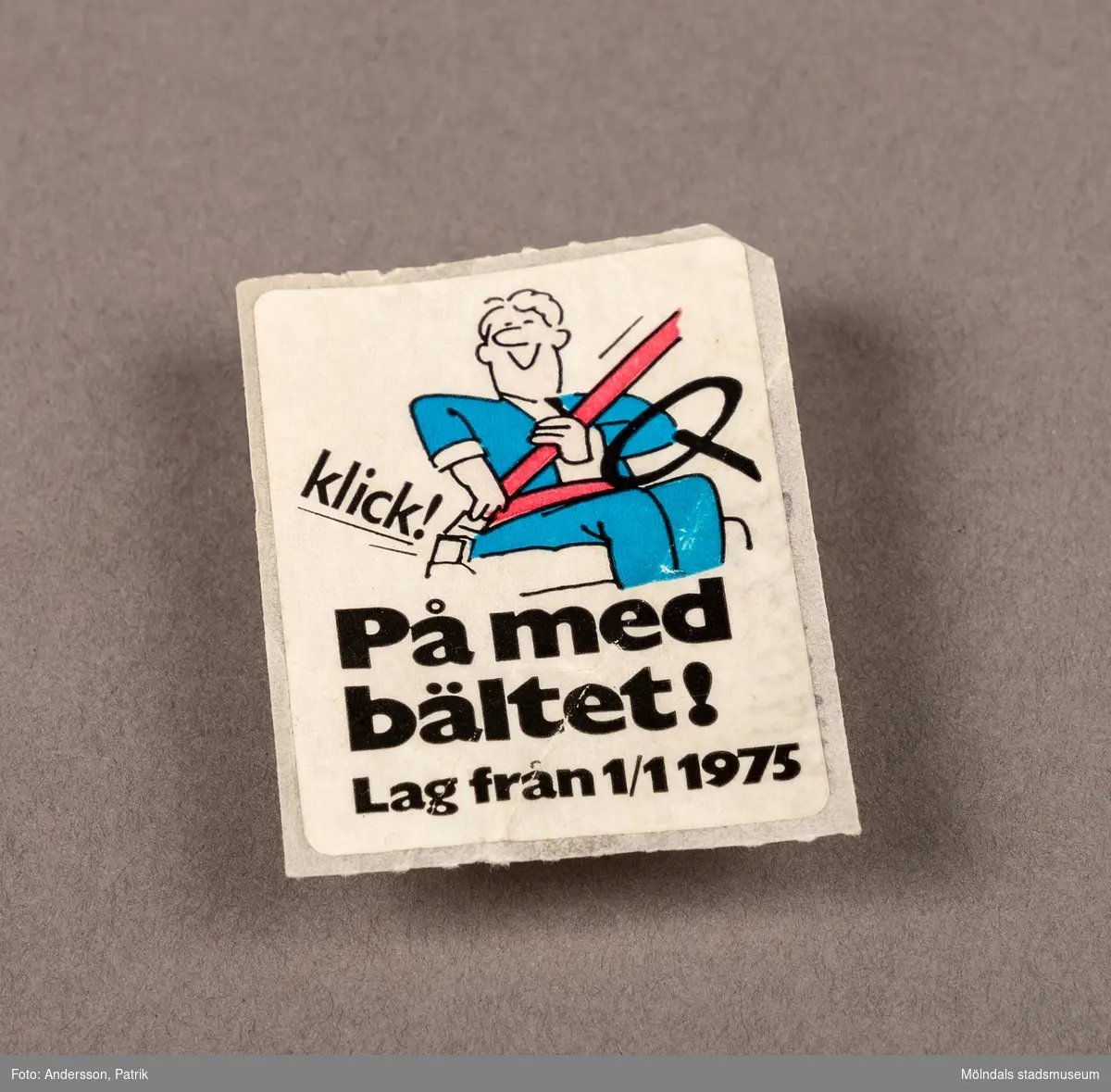 Klistermärke som föreställer en man som tar på sig ett bilbälte.

På märket står det: På med bältet! Lag från 1/1 1975.