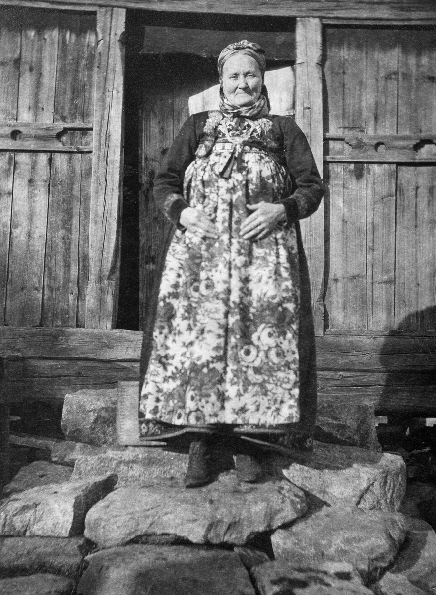Godt voksen kvinne fotografert på steintrapp foran inngangen til eldre våningshus.  Kvinna er iført tradisjonell stakk og hodekledning med lokalt særpreg fra Hallingdal.  Hun holder hendene på magen, som er dekt av et forkle med blomstrete mønster