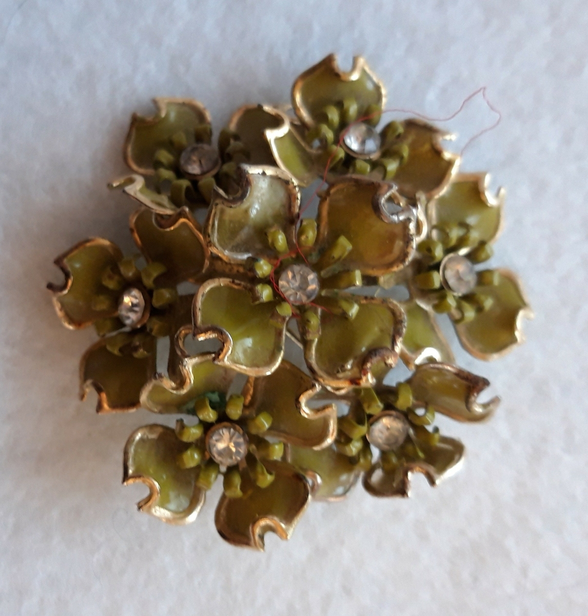 Nål i metall. Sju blomsterkroner er satt sammen og danner en rund nål. Kronbladene er lakkert i en gulgrønn farge. En klar sten er montert i midten av hver blomst.