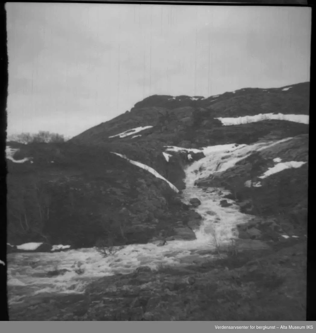 Bilde av elv med foss og noen snøflekker på et fjell.