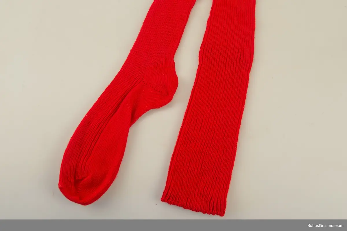 Del av bohusdräkt.
Enfärgad resårstickad röd strumpa handstickad av ullgarn, slätstickning under foten. 
För att hålla strumpan på plats ska möjligen ett strumpeband knytas över knäet.
Samhör med UM033950.