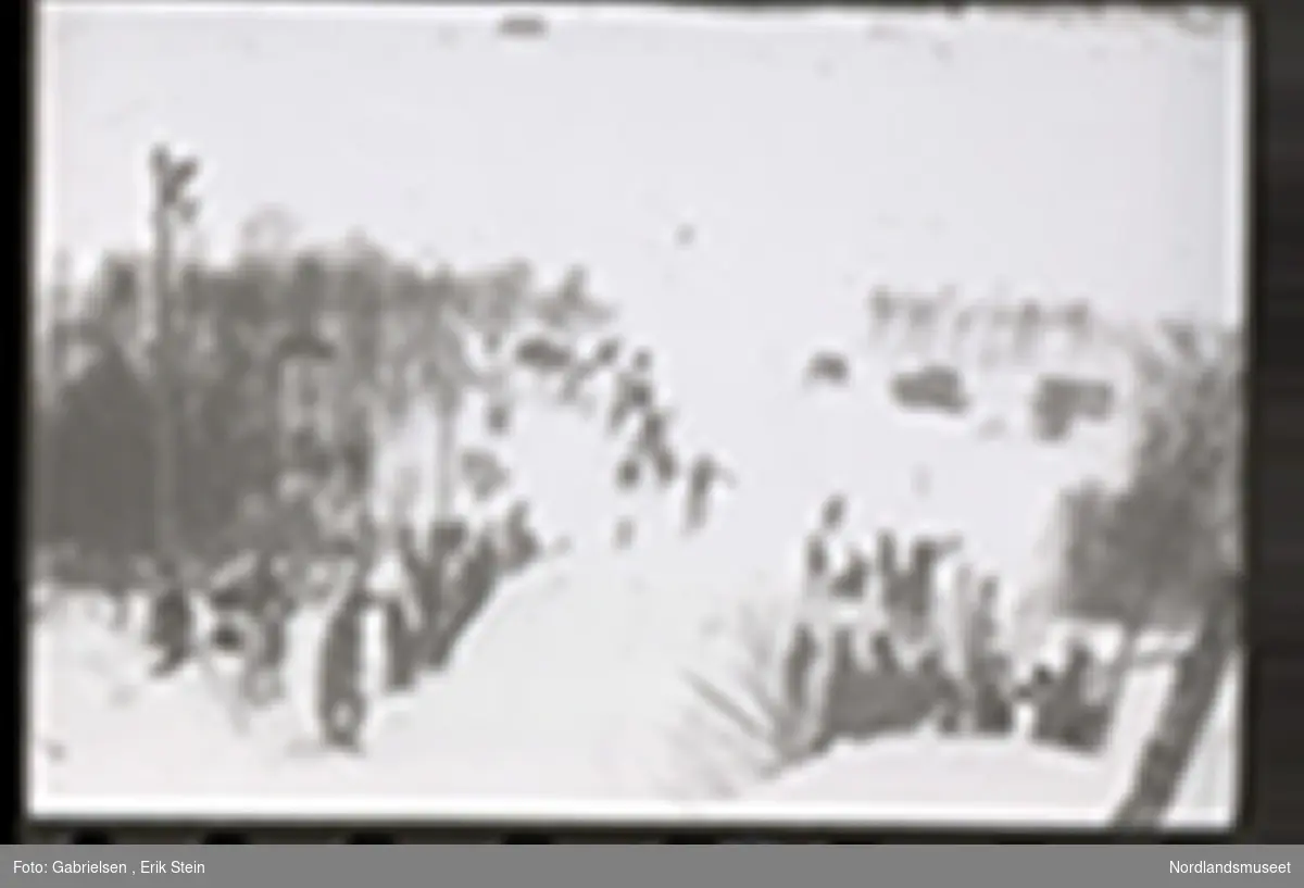 Fotografi av et skihopp som det hopper et barn med ski på seg fra mens noen kvinner og menn og barn med ski og skistaver og vinterklær og deltakernummer på seg som står og ser på barnet som hopper fra skihoppet vedsiden av skihoppet og lenger nede i bakken og tre barn som står i et utsiktstårn 
og ser på skihopperen og noen biler og busser som står parkert lenger nede i skibakken vedsiden av en vei som det vokser noen trær vedsiden av og man ser en skiløper ute i snømarken som har nettopp landet i snøen etter å ha hoppet fra skihoppet
