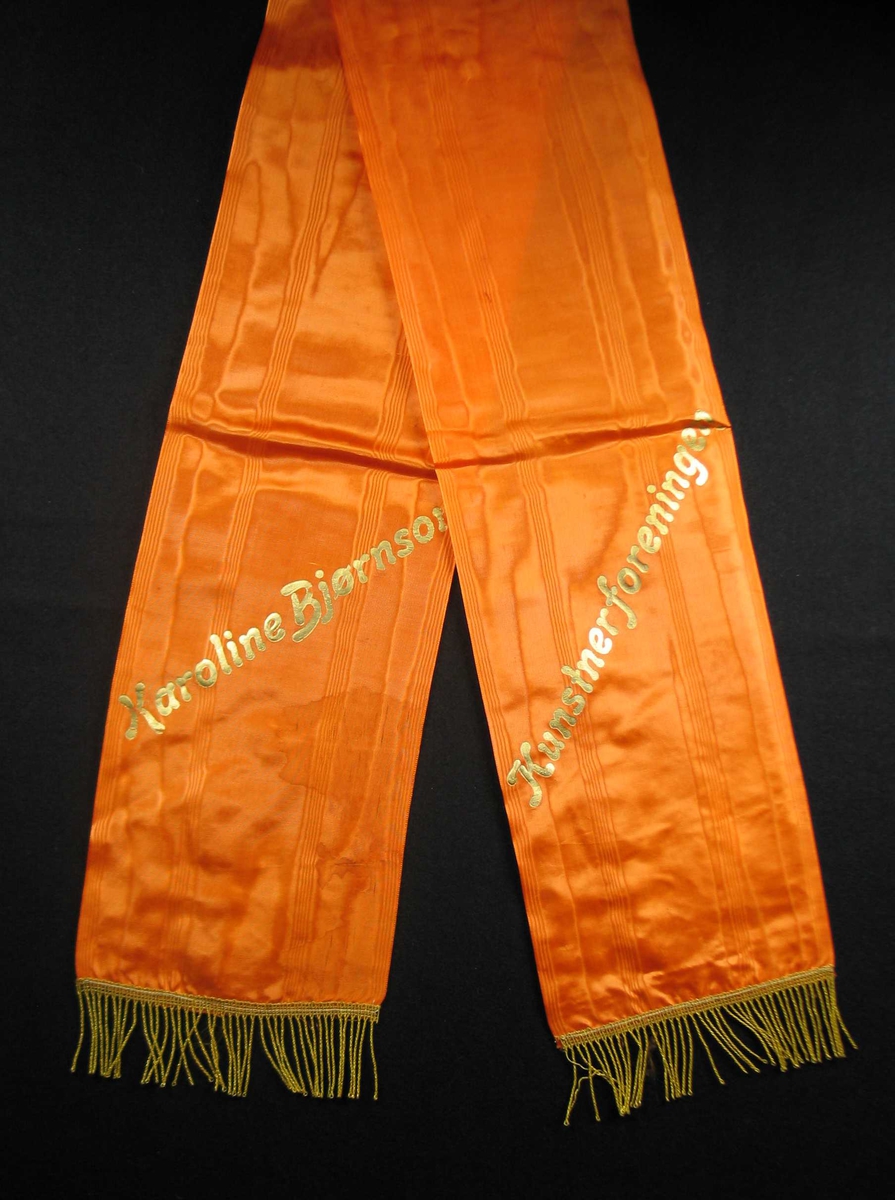 Begravelsessløyfe i oransje mønstret silke med tekst og frynser i gull.