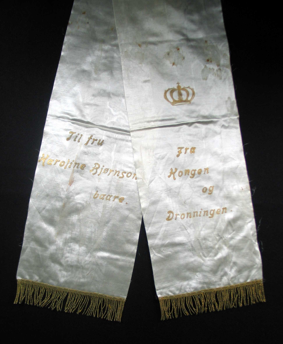 Begravelsessløyfe i hvit mønstret silke med tekst og frynser i gull.