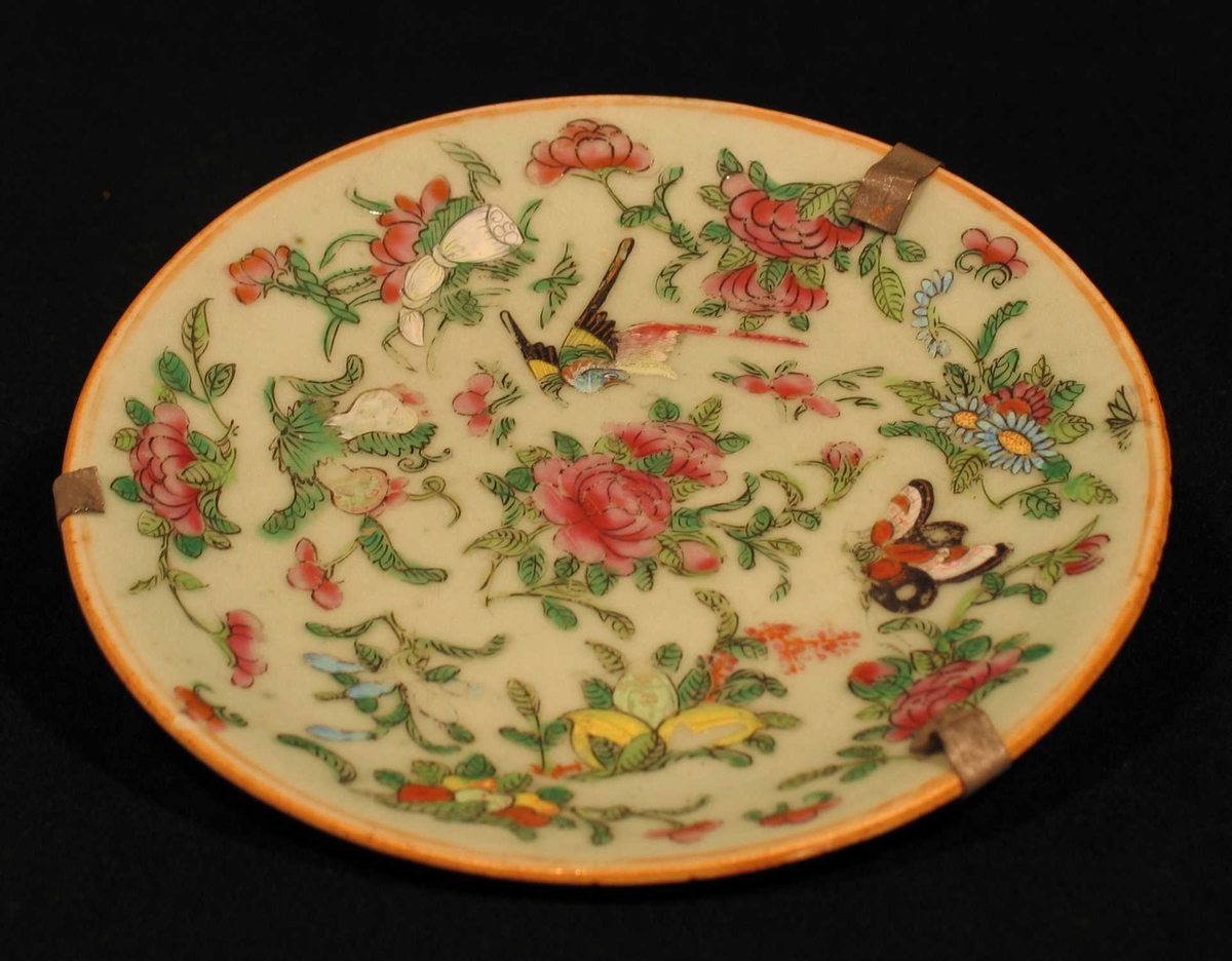 Asjett i porselen med lys grønn glasur og polykrom dekor. Motivet er blomster, sommerfugl og fugler. Oppheng på baksiden.