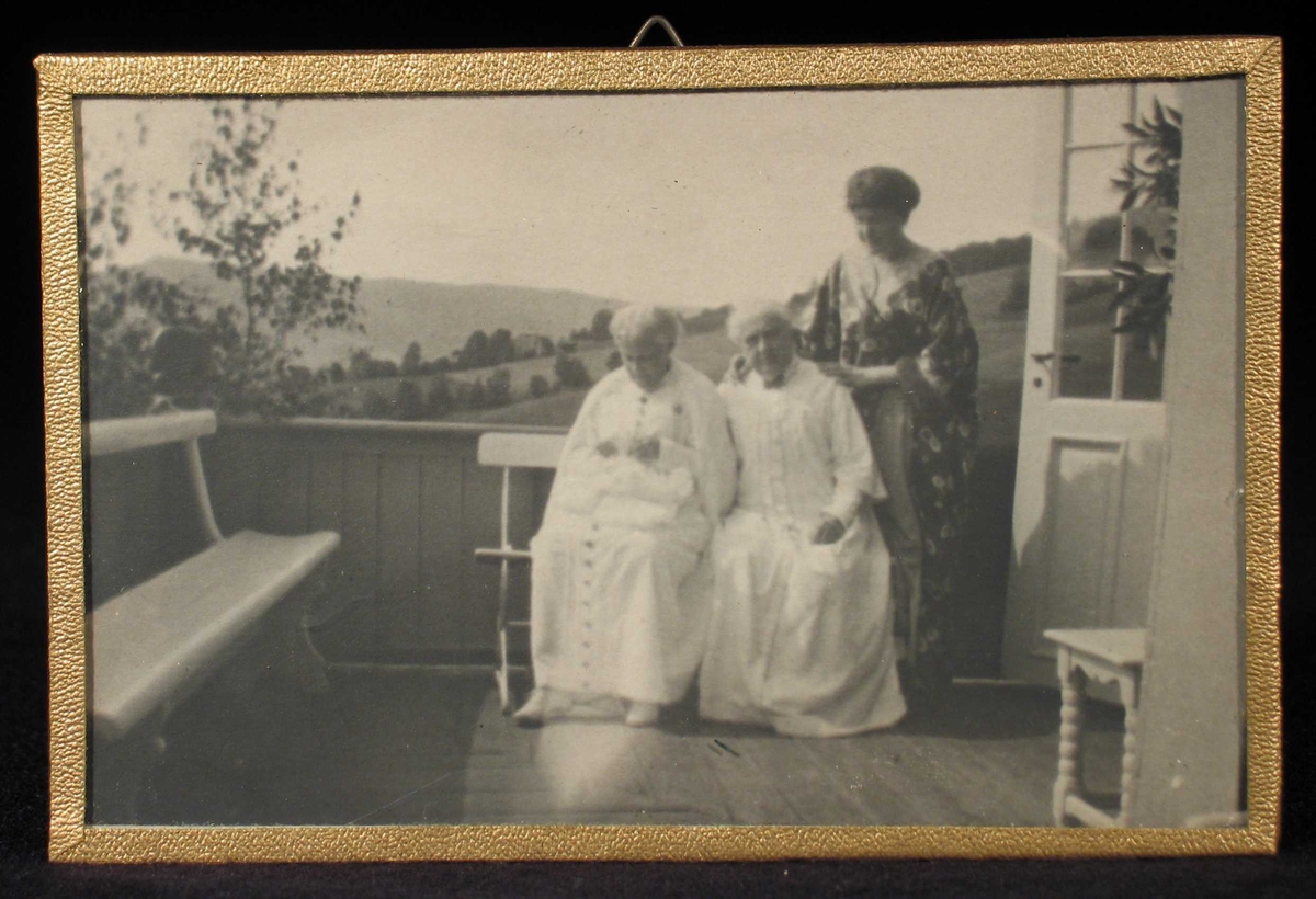 Tre kvinner på en veranda, to en benk og ei stående bak.