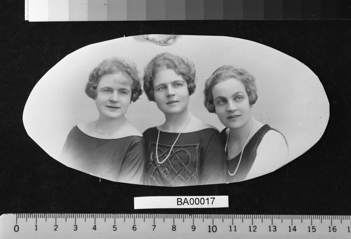 Atelierfotografi; familiebilde av tre unge damer i festkjoler, perlekjeder rundt halsen og fine korthårsfrisyrer lagt i bølger inn mot ansiktene.
Brystbilde i oval bredde-ramme.