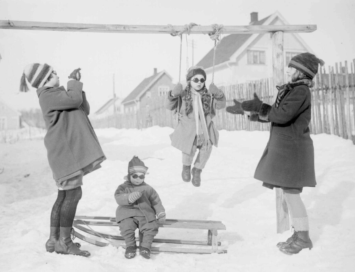 Vinter. Barn med snøbriller som leker i huske og med kjelke. Gerda Johansen.