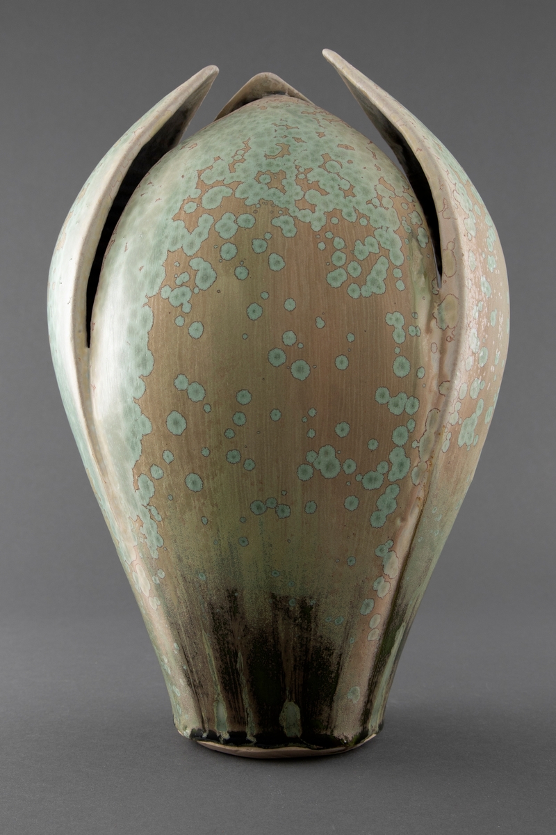 Håndlaget vase i delvis dreiet, delvis modellert steingods. Glasuren er beige i grunntonen med grønne flekker spredt over formen. Vasen er inspirert av naturens formspråk og ligner en knopp som er ved å springe ut.