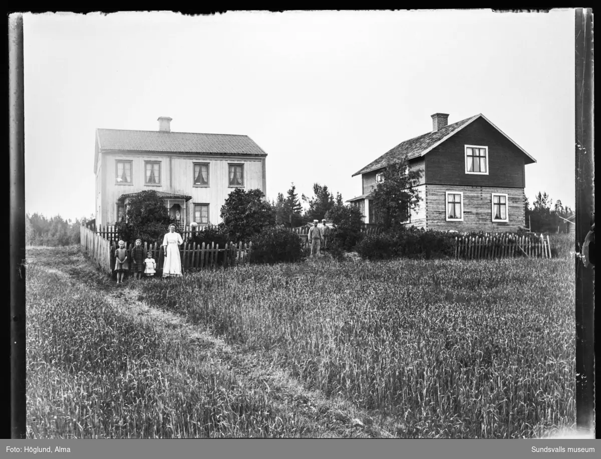 Två vyer mot gården Jöns Ols i Gräfte med två bostadshus och trädgården inhägnad med spjälstaket. Fyra barn, två kvinnor och två män syns på bilderna.