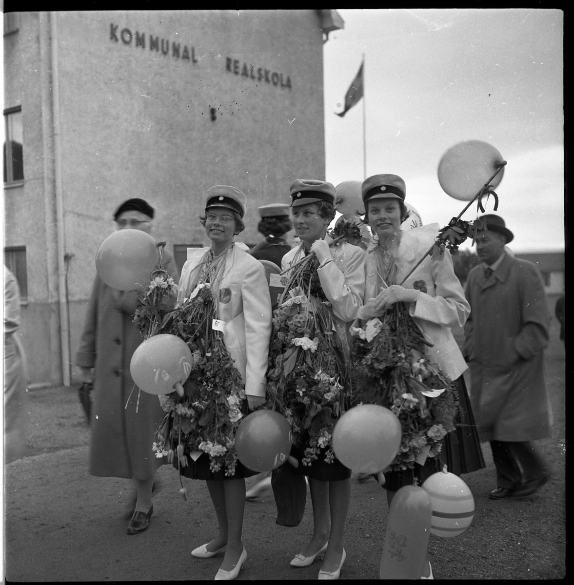Tre kvinnliga realister står uppställda för fotografering. De bär realskolemössor och de har buketter och ballonger runt halsarna. I bakgrunden syns skolbyggnaden, på fasaden står det kommunal realskola.