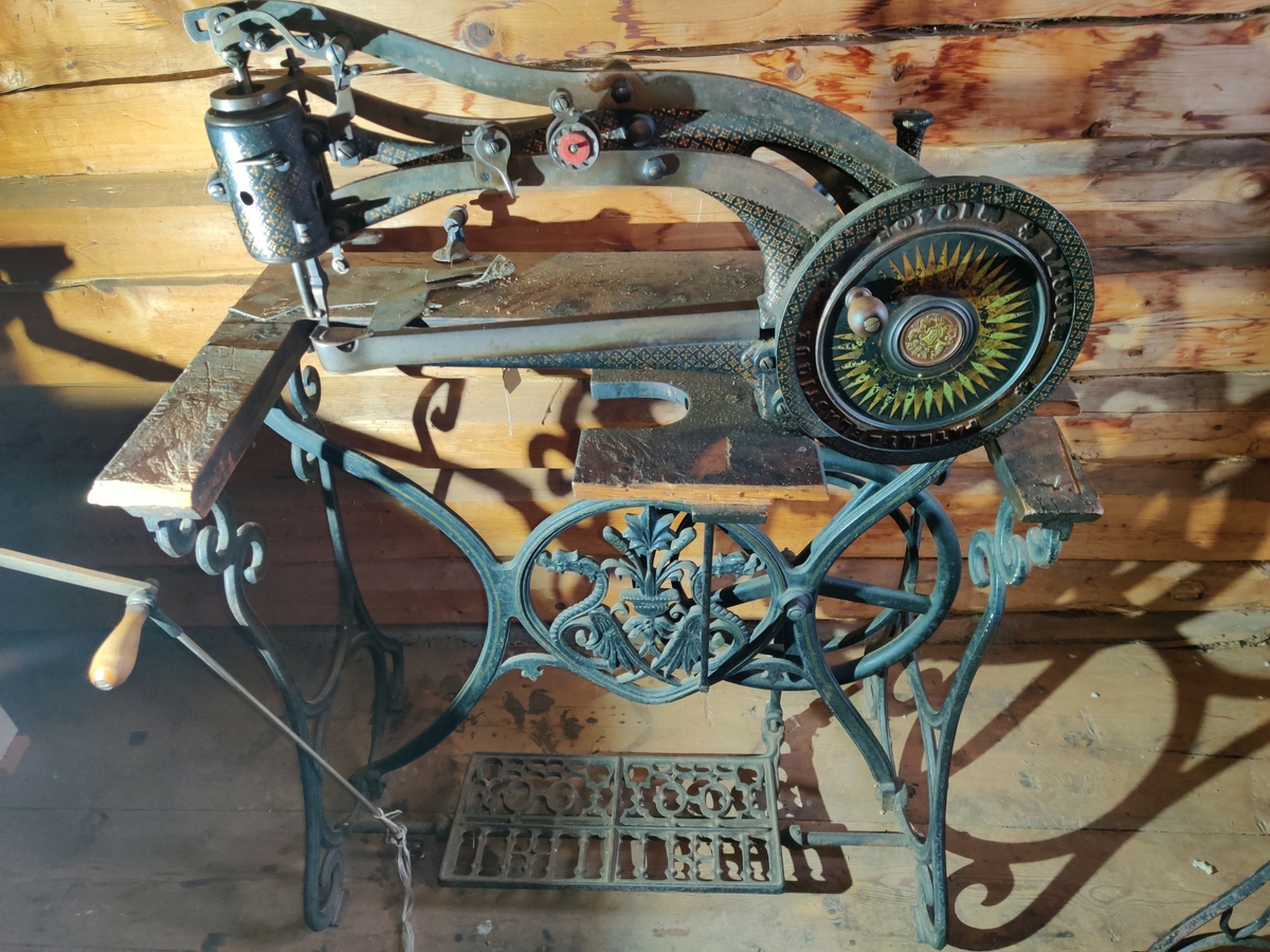 Symaskin for lær og skomakerarbeid, håndsveiv- og trådrevet, med bord av tre og på stativ av støpejern.