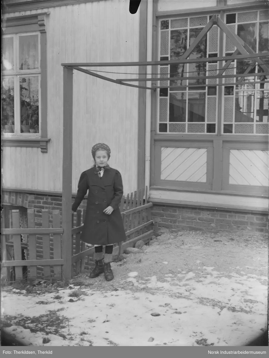 Jente kledd i kåpe og lue stående foran Solvang, senere kalt Hellandgården. Bygningen har dobbel inngangsdør og smårutete vinduer i deler av fasaden.