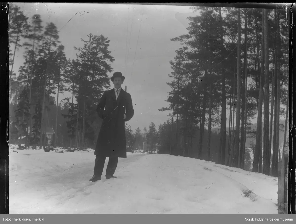 Mann kledd i hatt og frakk stående på gangvei ved boligområde.