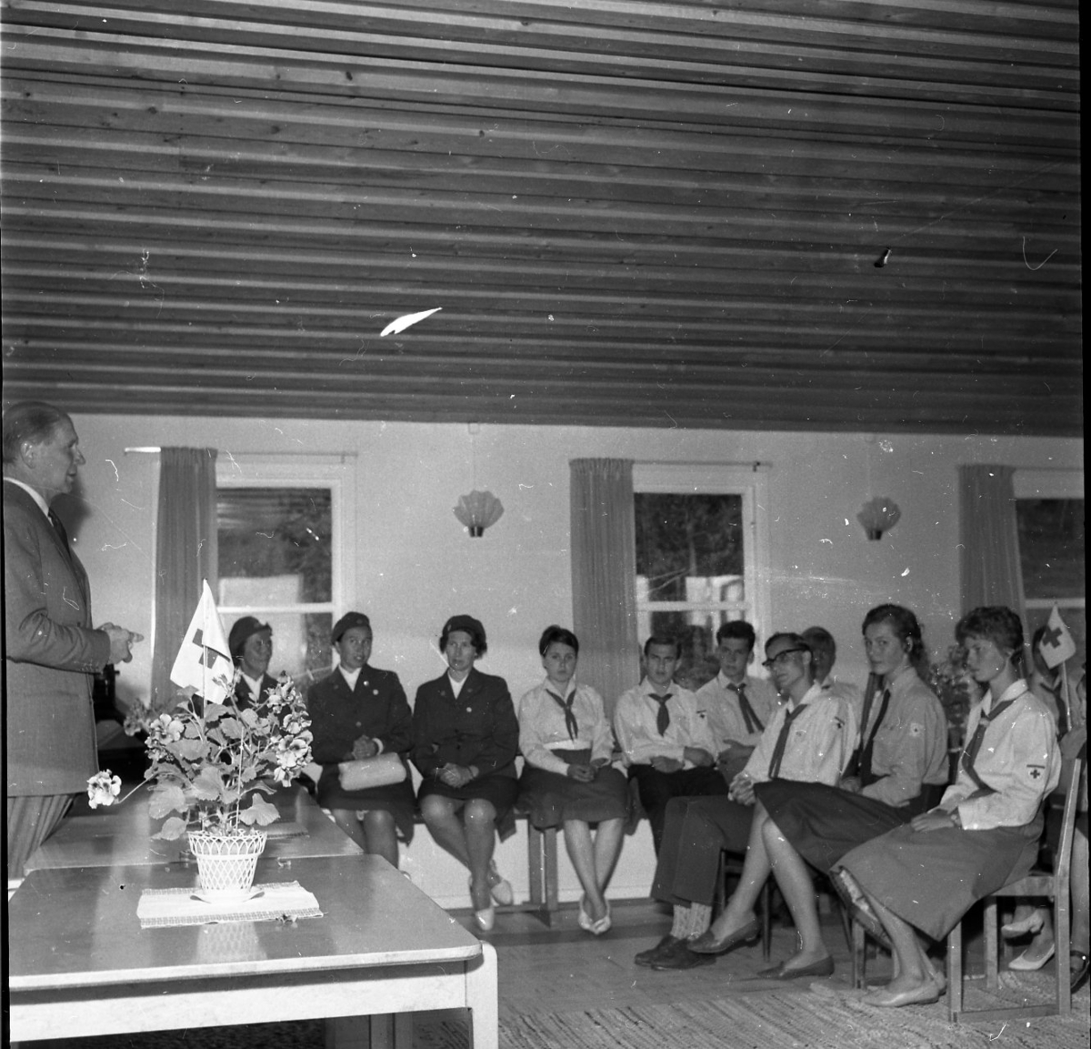 Röda korsläger i Ralingsås 1960-1961. Flera persoer i röda korsets kostymer sitter och lyssnar på en manlig talare. På ett bord står en krukväxt med en röda korset-flagga nedstucken.