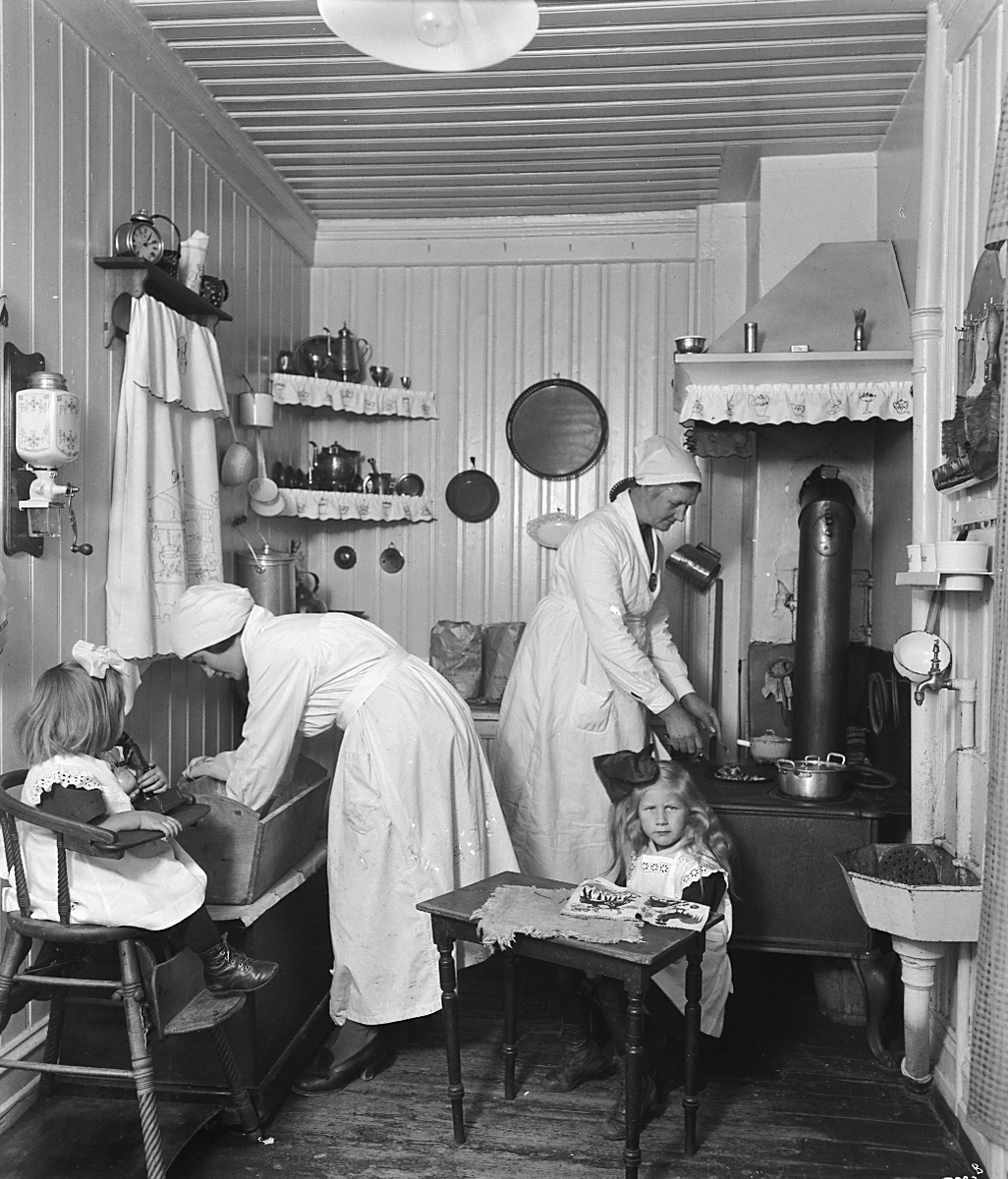 Hjemmekonsulenter steller på et kjøkken, to små piker sitter på stoler. Fotografert 1923.