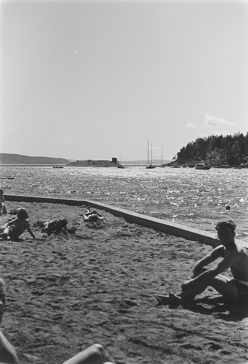 Solbading ved stranden med sjø og båter. Fotografert 1940.