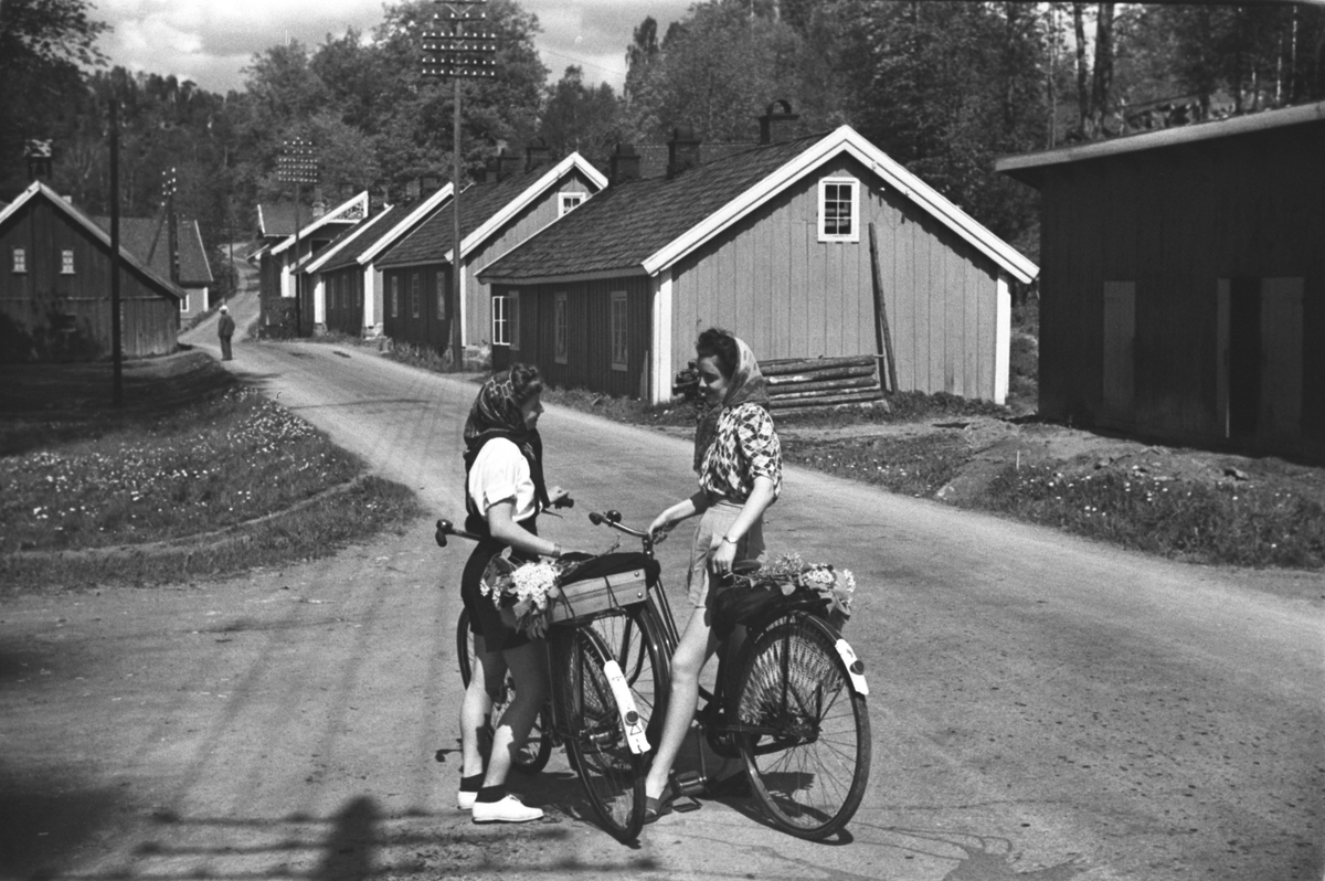 To kvinner på sykkeltur, her ved Bærum Jernverk. Langs veien ligger arbeiderboligene på rekke og rad.