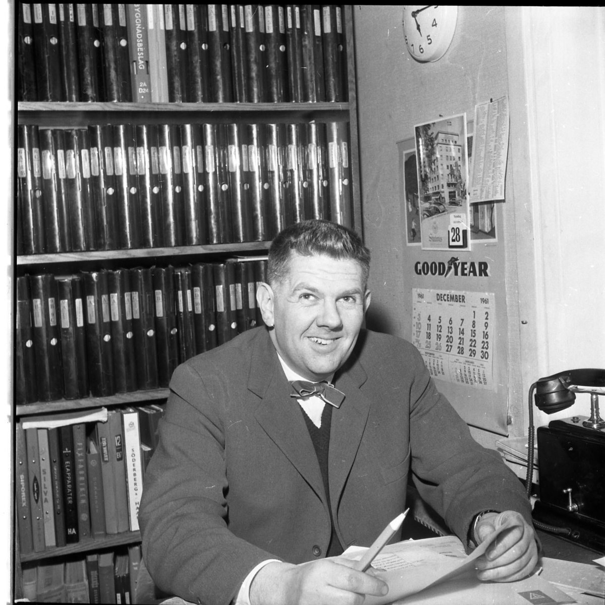 En man sitter på ett kontor. Bakom sig har han en bokhylla fylld med pärmar. Han ser glad ut. Det hänger en väggalmanacka på väggen och det står en telefon bredvid honom.
