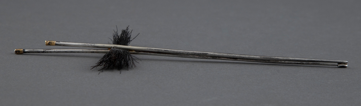Tre nåler som består av to blanke metallpinner av ulik lengde som er festet sammen. På tvers, mellom dem, er det en bunt svart hestetagl. Pinnene er forgylt på toppen.