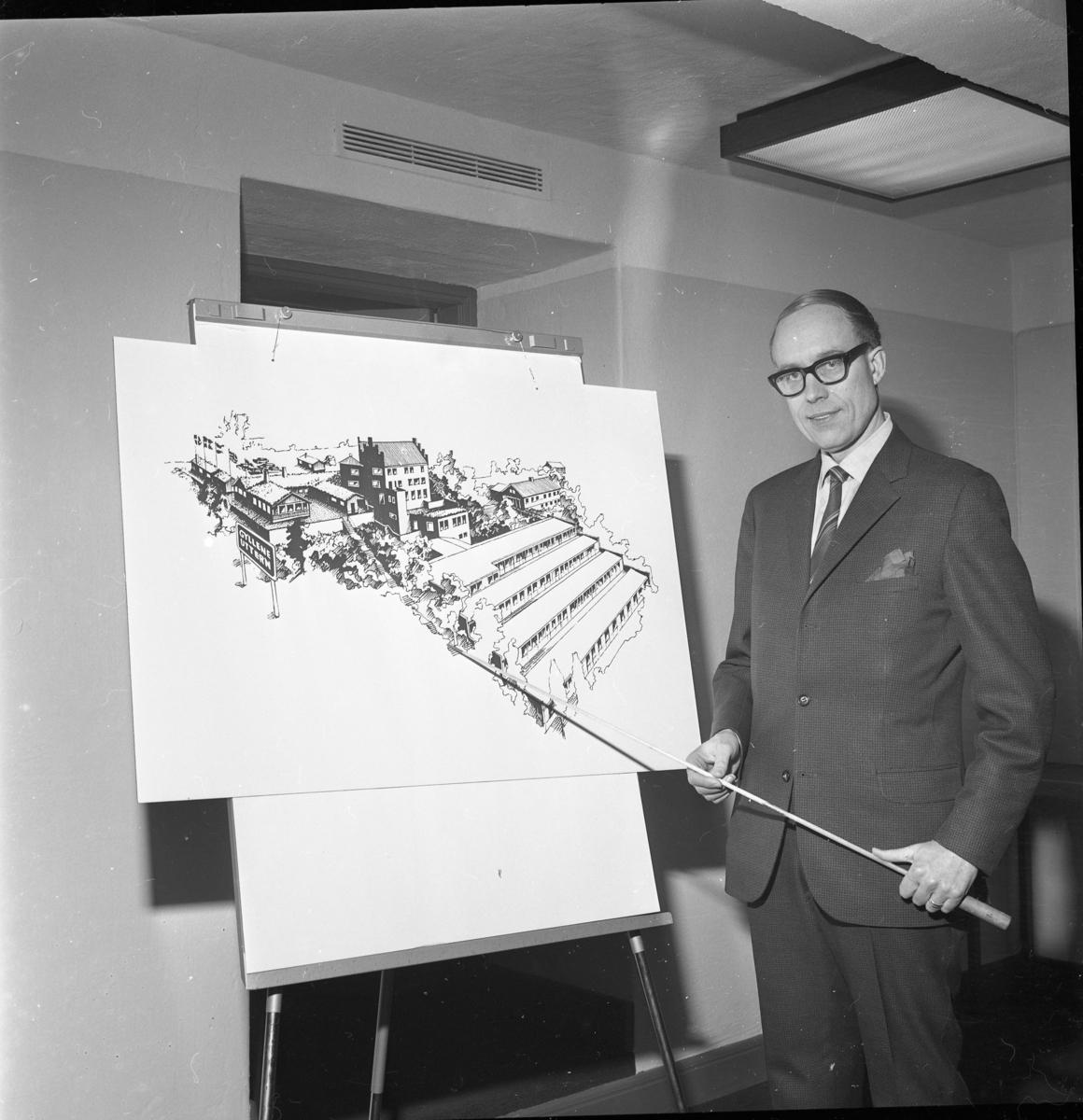 En man med glasögon, kostym och slips pekar med en käpp på en illustration av restaurang/hotell Gyllene Uttern efter tänkt tillbyggnad.