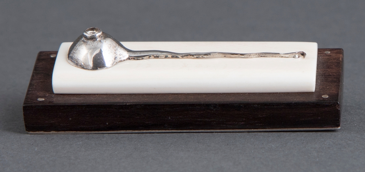 Brosje med rektangulær grunnform. Bunnnplate av ibenholt med en mindre plate i elfenben oppå. På denne er det en dekorasjon i sølv i form av et rumpetroll. Midt på denne er det nedfelt en briljant. På undersiden er det en plate i sølv med nål.