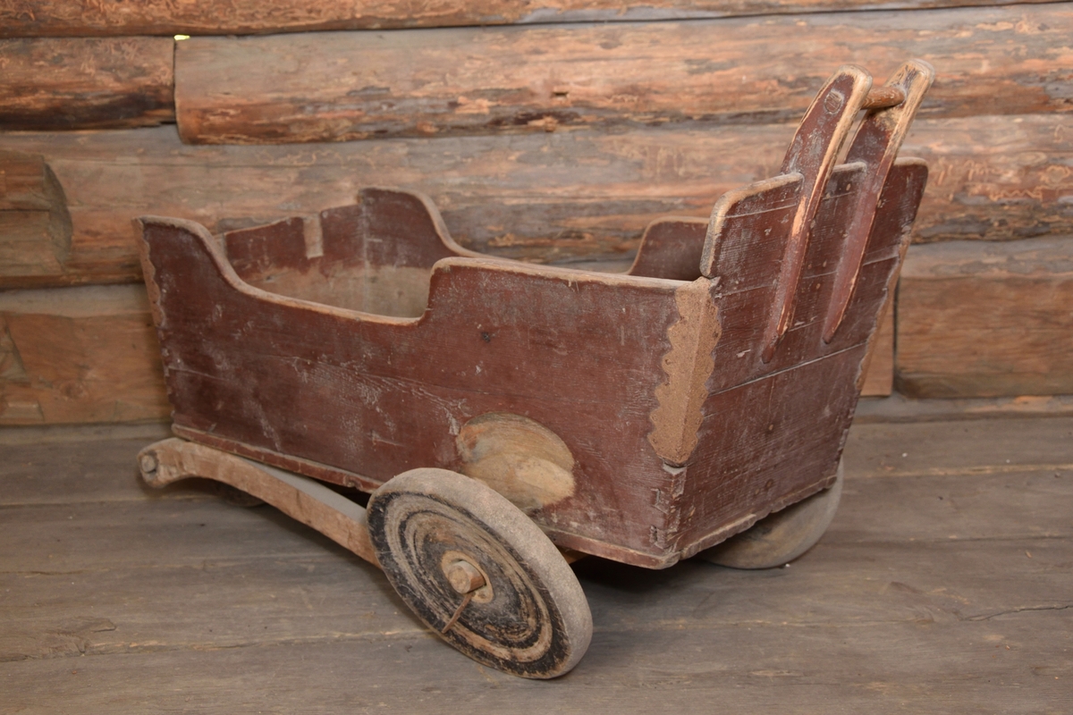 Barnvagn tillverkad i trä, rödmålad. Rektangulär form. Djup med raka sidor samt med ett handtag i bakkant. Barnvagnen med tre stycken hjul, två bak och ett fram.