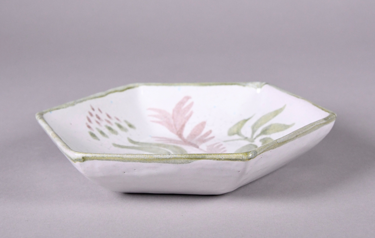 Skål av glassert keramikk, med opphøyd rand. Skålen er sekskantet, og har hvit grunnfarge. Håndmalt dekor av blomster og bladverk, og en grønn borde langs randen.