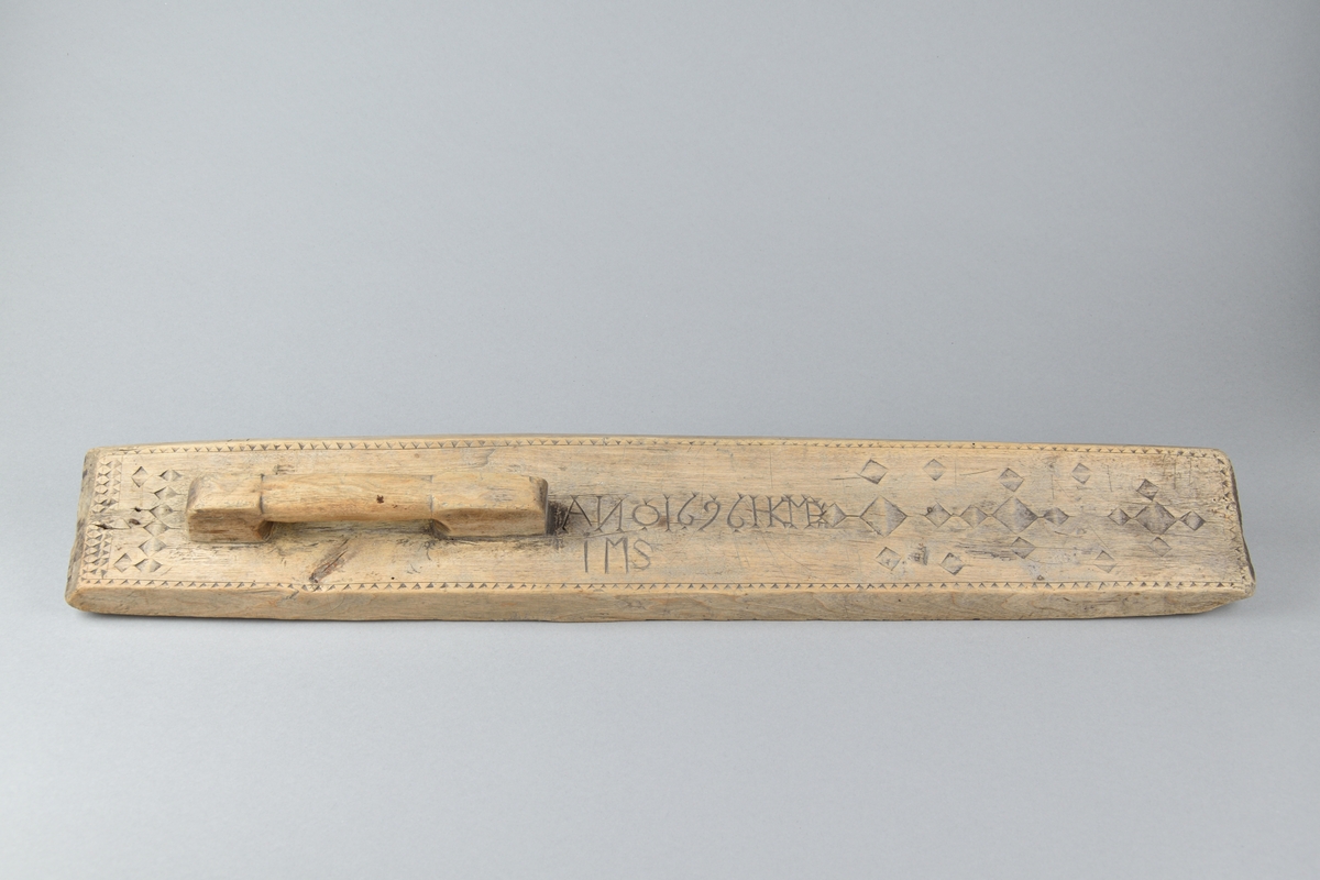 Kaveldon tillverkat i trä. Långt och fyrsidigt bräde med handtag. Ovansidan ornerad med romber och sicksack mönster, samt inskriften "ANO 1696 HKMDIMS".